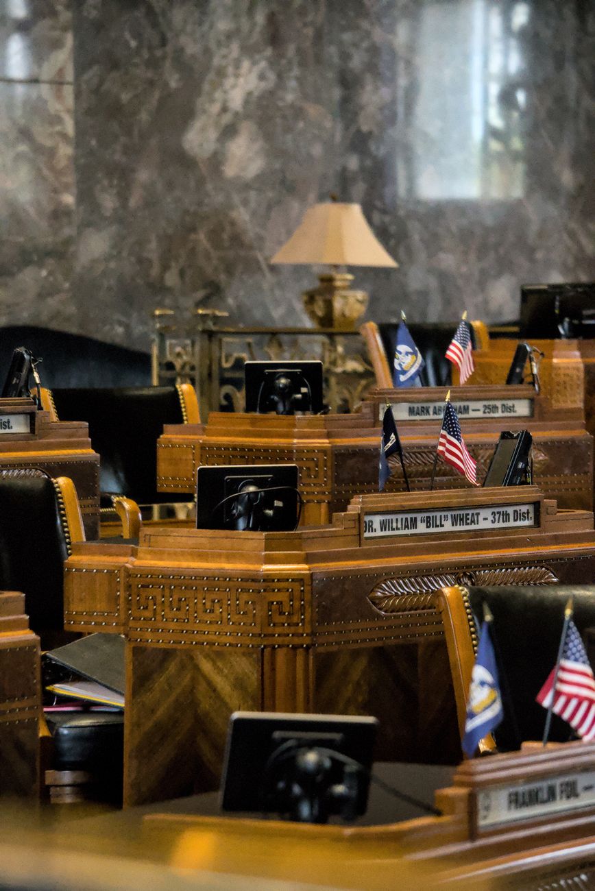 A legislative chamber with wooden desks, nameplates, and small American flags.
