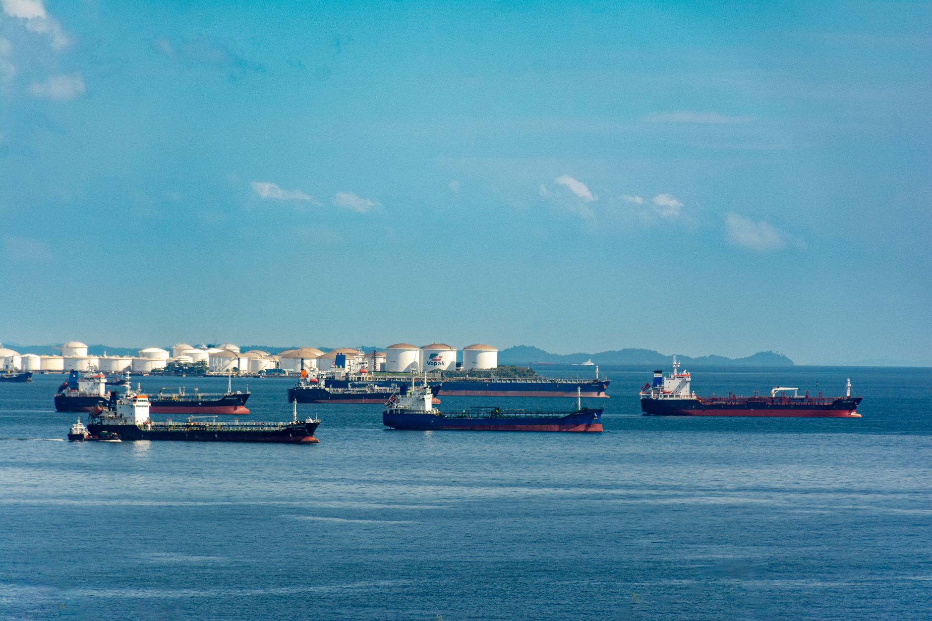 A fleet of cargo ships docked near oil storage tanks along a serene coastline with a clear blue sky above.