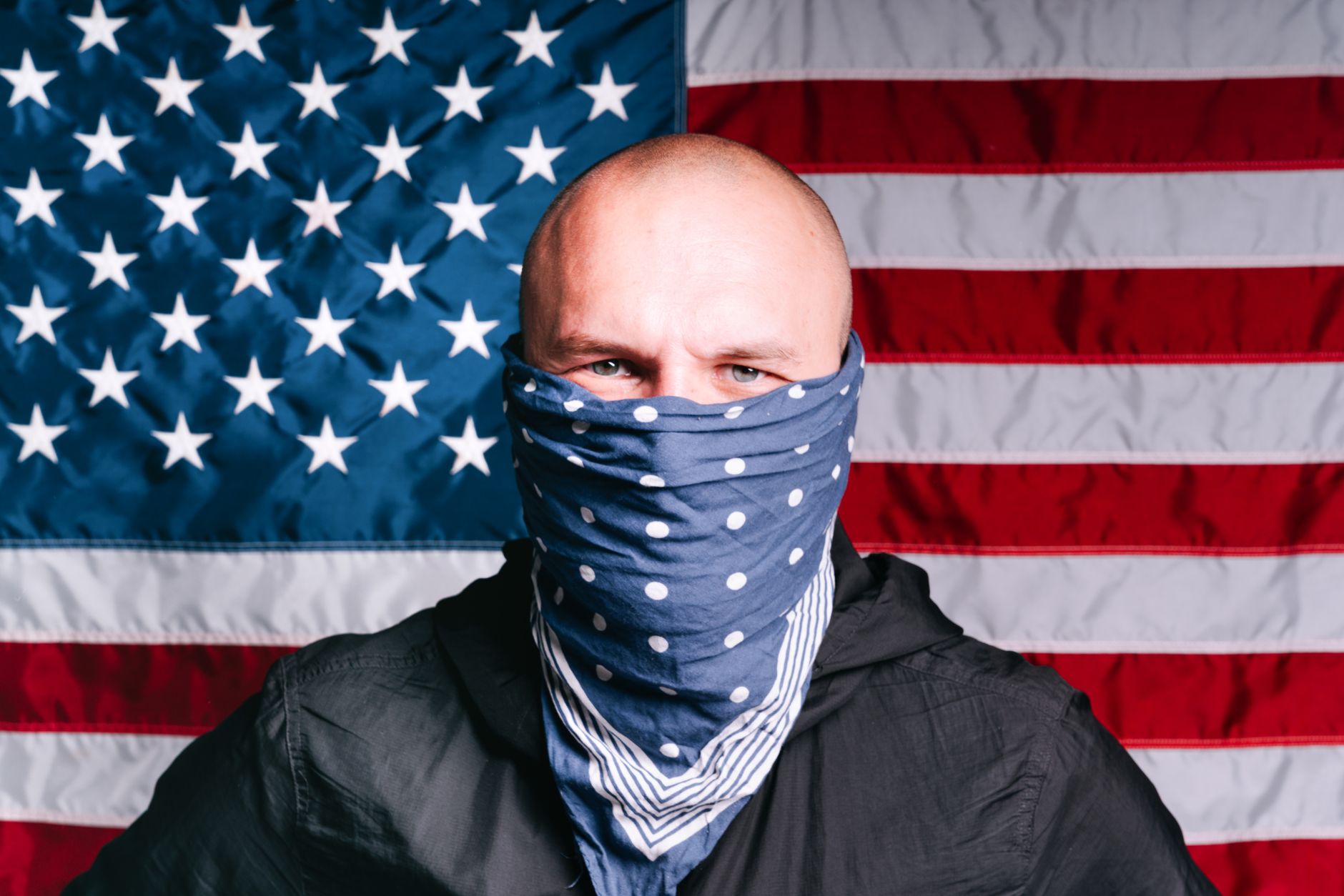 Bald man with face mask poses against American flag backdrop.