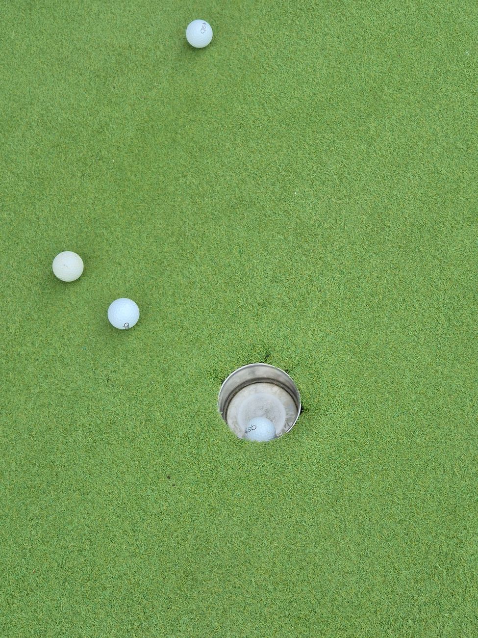 Mini golf balls on putting green around a hole, bright daylight.