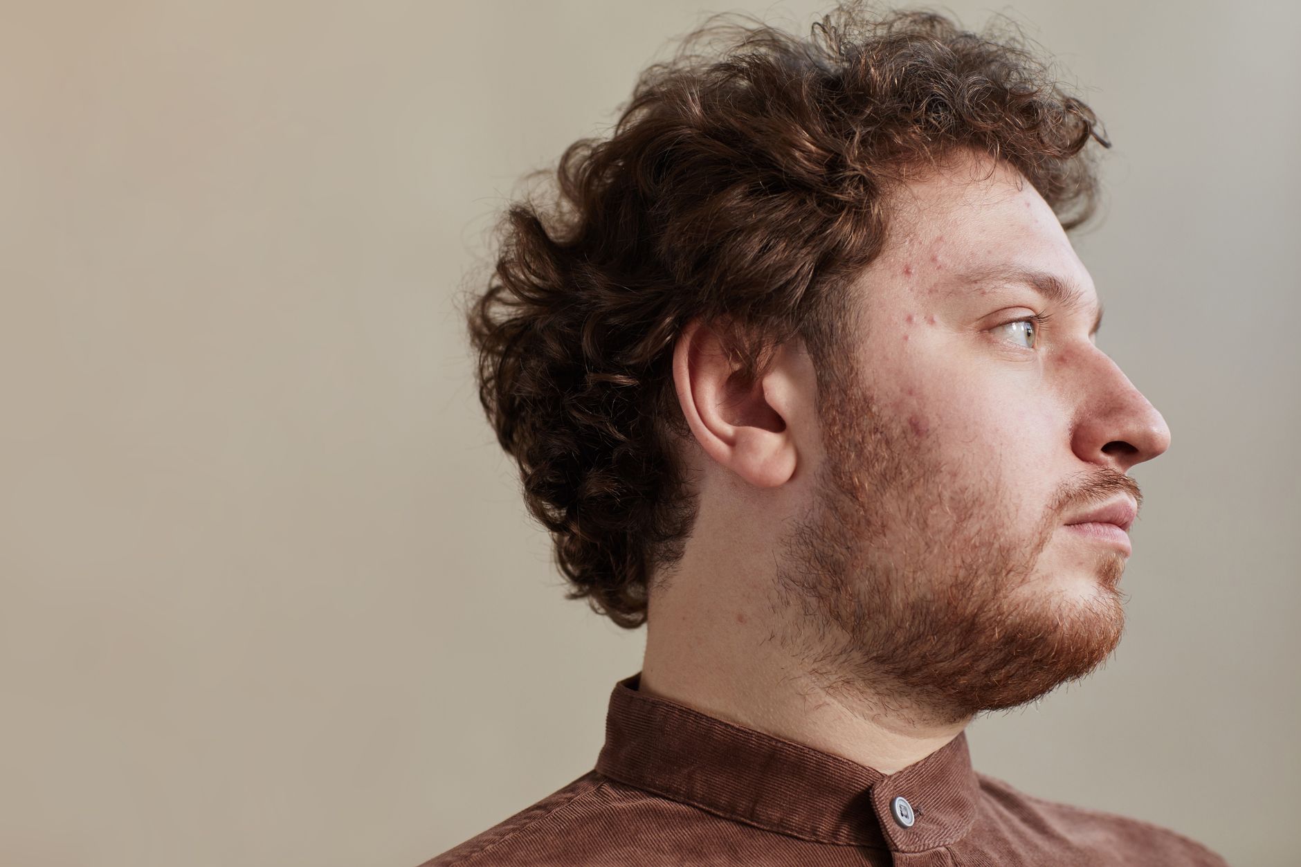 Close up profile of a serious man with curly hair and a beard indoors.