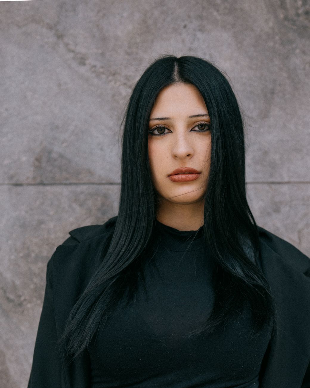 Stylish portrait of a woman with long black hair against a textured stone wall.