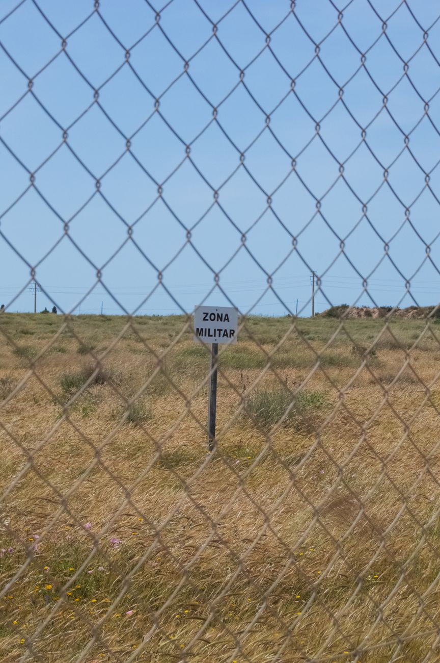 A military zone sign behind a chain link fence in a grassy field on a sunny day.