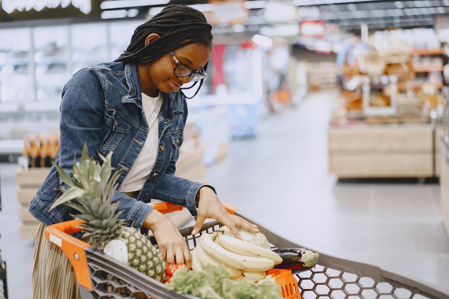 A woman shops for fresh fruits and vegetables in a supermarket, pushing a cart filled with groceries.