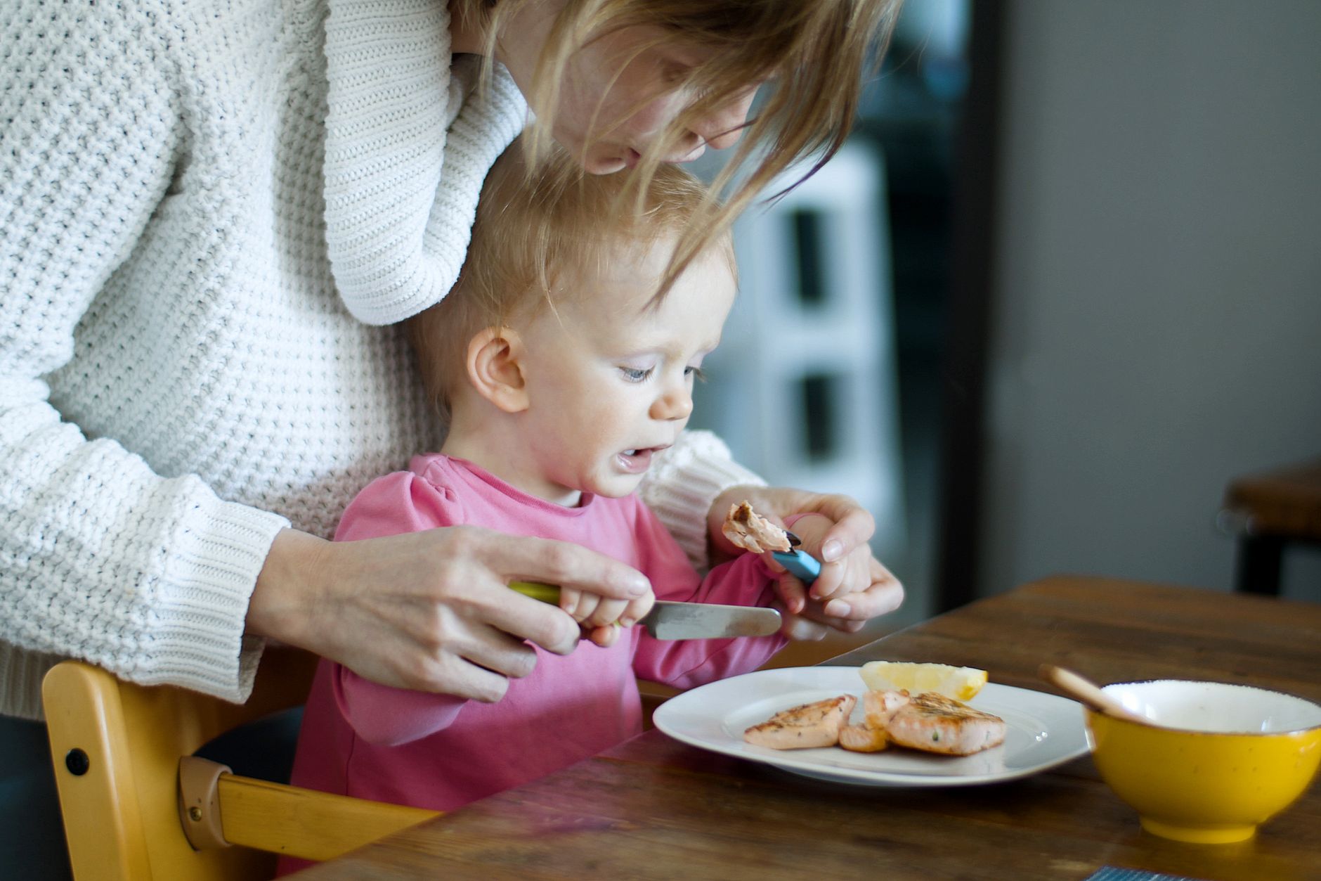 A mother assists her baby in eating salmon at breakfast, fostering bonding and nutrition.